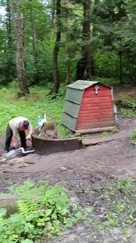 A person is filling a large plastic bottle with water from a tap located next to a circular stone structure. A small red structure with a green roof and a cross on top is visible behind the tap. The environment is a forest with trees and greenery. Another person is walking away from the tap, carrying two plastic bottles. A car is visible in the background on a dirt road.
