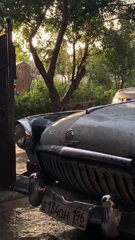 The video shows a close-up shot of the front of a vintage car. The car is parked outdoors, with trees and foliage in the background. The sun is setting, casting a warm glow on the scene. The car appears to be old and possibly weathered, with some dust and water marks on its surface. The license plate is visible and reads E1340H 196. There is a gate on the left side of the frame. The overall impression is one of nostalgia and a quiet, peaceful atmosphere.