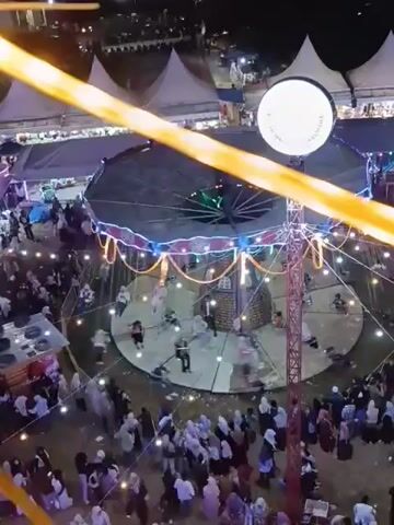 This video provides an aerial view of an outdoor night market or festival. A central amusement ride, possibly a carousel or swing ride, is operational with people riding it. The ride is illuminated with colorful lights and surrounded by tents and stalls. A crowd of people is gathered around the ride and navigating through the market area. The scene is lit by artificial lights, including string lights and spotlights, creating a festive atmosphere. The overall impression is that of a lively and bustling event at night.