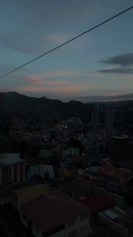 The view from inside a cable car in La Paz, Bolivia, reveals a city nestled among hills under a dusky sky. The sky is a mix of dark blue and soft pink hues as the sun sets or rises. Illuminated streets wind through the city, contrasting with the dark outlines of buildings and the looming hills. The cable car's infrastructure, represented by a single, taut wire, stretches across the top of the frame. The foreground shows a collection of rooftops.