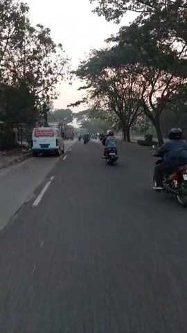 This is a first-person perspective video from a moving vehicle, likely a motorcycle, on a road. Several other motorcycles with riders are traveling in the same direction. On the left side of the road, there is a white van with "LALAMOVE" printed on it, parked alongside other businesses. The road is paved and has white lane markings. Trees line the right side of the road, and the sky appears hazy or overcast, possibly indicating sunrise or sunset. The overall atmosphere is that of daily commute or transport in an urban or semi-urban environment.