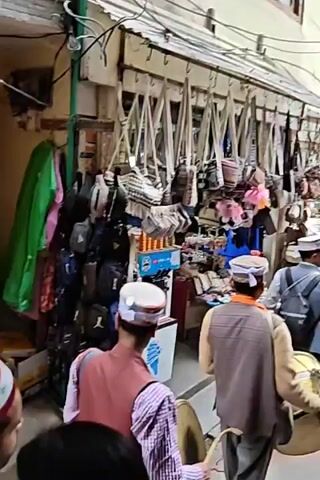 The video captures a lively scene within a narrow, bustling market alley in Kasol, India. The setting is an outdoor commercial area, with market stalls packed closely on both sides of the pathway.

The primary subjects are a group of men moving through the alley, many of whom are wearing traditional Himachali caps (topis) with colorful bands. One man is prominently featured playing a large Dhol drum, while another plays a wind instrument, possibly a Shehnai or similar, creating a distinct musical sound. These musicians appear to be part of a cultural procession or performance, as they walk together through the crowded space. The man playing the wind instrument wears a light brown vest over a checkered shirt, and the drummer also sports traditional headwear.

The market stalls are laden with merchandise. On the left side, various woven and fabric bags, including sling bags and small purses, are stacked and displayed. On the right, numerous bags of different styles, colors, and materials hang from overhead racks, along with shelves displaying smaller items like colorful trinkets, toys, and souvenirs. Green and pink fabrics are also seen hanging in the background, adding to the colorful array of goods.

The activity involves the musical procession navigating through the market, with other people, likely shoppers, moving around and observing. The atmosphere is vibrant and culturally rich, characterized by the traditional music and the busy market environment. The video appears to be shot during the daytime, with sufficient natural light illuminating the scene. No discernible English or local language text is legible on any signs or products.