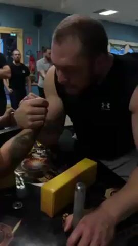Here is a description of the video:

The video shows a man arm wrestling another man. The man on the right is wearing a black Under Armour sleeveless shirt and grey pants. He has a beard and short brown hair. The man on the left is wearing a black t-shirt and a baseball cap. He has a tattoo on his left forearm. They are arm wrestling on a table with a yellow block in the middle. There are other people in the background watching them.