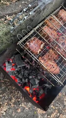 A grilling basket filled with pieces of meat is positioned over glowing coals in a charcoal grill. The meat appears to be chicken or pork, with some pieces showing grill marks. The scene is outdoors, with foliage and debris visible in the background. The camera angle is from above, looking down at the grill. The video is brief, lasting only a few seconds, and shows the meat sizzling and cooking.