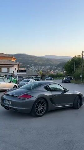 The video captures a street scene with several cars parked and moving, set against a backdrop of rolling hills and a town. In the foreground, a dark gray Porsche Cayman is the most prominent vehicle, facing away from the camera. The car has a Bulgarian license plate that reads CB 2133 BE. To the left of the Porsche, a silver sedan is parked, and further back, a black car and a silver sedan are visible on the road. In the distance, a town with buildings is nestled among green, tree-covered hills, which are bathed in the soft light of what appears to be late afternoon or early evening. The sky is clear and a pale blue. There are no people visible in the video. The overall impression is one of a quiet, scenic residential area.