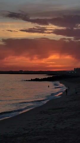A video captured at dusk shows a serene beach scene. The sky is ablaze with vibrant hues of orange and red, painted with dramatic clouds. The gentle waves of the ocean softly lap onto the sandy shore, creating a tranquil atmosphere. In the distance, a lone figure can be seen wading in the shallow water, while closer to the shore, two people are standing on the beach, appearing to observe the sunset. The overall ambiance is one of peacefulness and natural beauty as the day gracefully transitions into night.