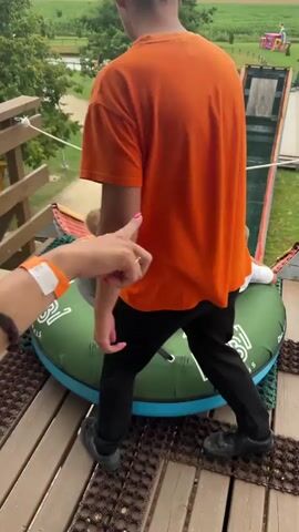 The video shows a group of children preparing to go down a steep slide on inflatable tubes. In the foreground, an adult wearing an orange shirt stands to the left, facing away from the camera and pointing towards the slide. In the center, three young boys are sitting in a large green inflatable tube, positioned at the top of a wooden ramp that leads to the slide. The boys are facing away from the camera, looking down the slide. The slide itself is a long, green, downward-sloping surface with a textured matting. Several other inflatable tubes are visible on the ramp further down. In the background, there is a grassy area with trees, a pond, and a path. A girl is visible walking up the ramp in the distance. The overall environment appears to be an outdoor recreational park.