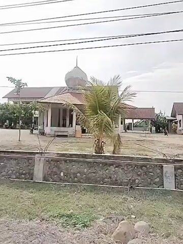 The video, filmed from a moving vehicle, shows a building with a dome and several structures with thatched roofs, likely a mosque, and surrounding landscaping. The camera pans to the side, showing trees and power lines, and then captures a view of the building complex again. The sky is overcast. An audio track with a religious chant is playing.