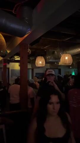 A crowd of people are gathered in a dimly lit bar or club. The ceiling has exposed pipes and ductwork, with several woven light fixtures hanging down. In the foreground, a young man in a dark t-shirt and a baseball cap is drinking from a bottle. Other people are visible in the background, some dancing and socializing. The atmosphere appears lively and crowded.