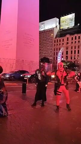 The video, shot at night, features three people in superhero costumes: Spider-Man, Venom, and Deadpool. They are standing on a brick-paved area in front of a large, brightly lit obelisk in Buenos Aires, Argentina. Traffic and city buildings are visible in the background. The Spider-Man and Deadpool characters take center stage, performing a coordinated dance routine, while Venom stands slightly behind them. The area is lit with a strong pink or reddish hue, probably from the obelisk's lighting. The overall impression is of street performers entertaining an audience or filming content.