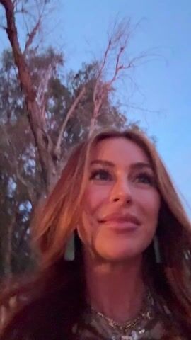 The video shows a woman outdoors as she is filming herself. She is smiling and talking about how the sun is rising, and that she has been partying all night. Behind her, a pink sunrise is visible through the trees. She is wearing turquoise earrings, a chain necklace, and a shirt with an animal print. Another person, whose face is obscured by the darkness, is walking beside her.