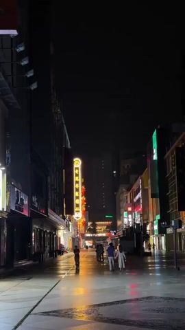 The video shows a street scene at night. The street is mostly empty with a few pedestrians walking. The ground appears wet, reflecting the bright lights from the buildings. Many of the buildings have neon signs in Chinese characters. The sky is dark and not much of it is visible. The scene is quiet and peaceful.