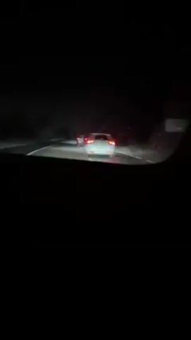 The video shows a car traveling on a highway at night in the rain. The view is from inside the vehicle, looking out through the windshield. Two other cars are visible ahead, their taillights illuminated in the darkness. The highway appears wet due to the rain, and the visibility is limited. The scene is somewhat dark and blurry, but the headlights and taillights of the cars provide some illumination.