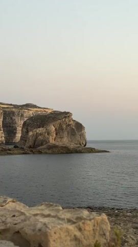 A vertical, slightly shaky video captures a rocky coastline meeting a calm, dark body of water. In the foreground, large, light-colored rocks are partially visible, with hints of green foliage. The midground features a prominent, weathered rock formation jutting out into the sea, its surface textured with layers of sediment and erosion. To the left, a larger, layered cliff face rises from the water. The sky above is a pale, cloudless gradient, transitioning from a light pinkish hue near the horizon to a pale gray at the top. The overall scene suggests an outdoor, natural environment, likely at dusk or dawn due to the soft, diffused light. No people, animals, or man-made structures are evident in the frame.