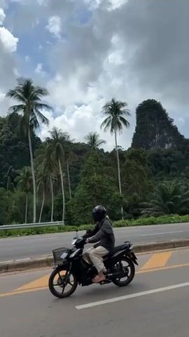 The video shows a view from a moving vehicle on a road. In the foreground, a person on a motorcycle is visible on the left side of the road. The road itself is paved with gray asphalt and has yellow and white markings. On the right side of the road, there are palm trees and lush green vegetation. In the background, there are large, dark, tree-covered hills or mountains under a partly cloudy sky. The camera appears to be moving forward, capturing the passing scenery. There is a street light visible on the right side of the road. The video also shows a silver car driving on the opposite side of the road. The overall environment appears to be tropical or subtropical.