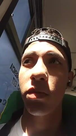 A close-up, eye-level shot shows a young man with short, curly hair, light skin, and a slight stubble, wearing a black baseball cap. He is wearing a white shirt and a jacket with a green and yellow stripe on the shoulder. The man is looking slightly upward at the beginning of the video, then turns his eyes to look directly at the camera. He is in a vehicle, most likely a bus. A window is visible in the background, along with a partial safety sign in reverse.