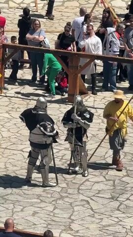 The video captures a scene with two individuals dressed in full medieval armor, standing within a fenced area paved with stone tiles. Each is holding a long polearm. They are surrounded by a diverse group of onlookers, including adults and children, some seated on the ground, others standing and observing. A wooden structure and rope form the enclosure, and the setting appears to be a sunny outdoor event or festival.