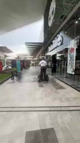 A man in a white shirt and dark pants is riding a small, four-wheeled vehicle down a paved walkway. The walkway is lined with stores, including a Timberland store with a green wall feature and window displays. There are benches and other people visible in the background. The sky is overcast. The camera is stationary and slightly low, looking up at the scene.