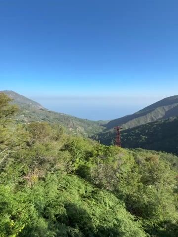 The video shows a mountain landscape under a clear blue sky. Lush green vegetation covers the foreground, with glimpses of houses nestled in the valley. A red metal tower stands prominently amidst the greenery, appearing to be a part of a cable car or ski lift system. In the distance, other similar towers can be seen against the backdrop of rolling hills, the ocean barely visible in the background haze. A bird is perched atop one of the towers. The camera zooms in to a medium shot on one of the red metal towers.