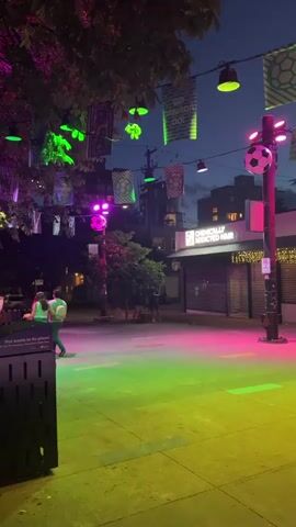 The video shows an outdoor urban scene at dusk. The ground is illuminated with multi-colored lights, transitioning from green to pink and then to purple. String lights and hanging banners, some with soccer ball designs and the text "GO CANADA GO!", are visible overhead. Several large, inflatable soccer balls are suspended in the air, along with smaller ones. A lamppost with soccer ball decorations stands prominently. In the background, there are buildings, including one with a sign that reads "CHINCHILLY ADDED HAIR." A few people are present in the scene, with some sitting and others walking. One person is sitting alone on a bench on the left side of the frame, and a group of people is visible in the middle ground. The overall atmosphere appears festive and suggests a celebration related to soccer.