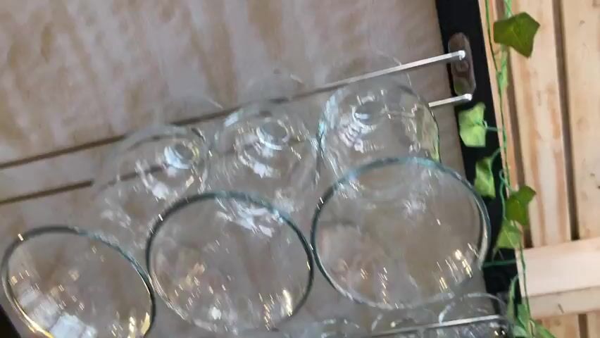 The video shows a close-up, angled view of a wine rack with multiple wine glasses suspended upside down from metal racks. The glasses are clear and have a spherical bowl shape. Behind the wine glasses, a shelf holds several bottles of wine, some with white labels and some with dark labels. Artificial green ivy is draped along the edge of the shelf, adding a decorative element. The background features wooden planks and a glimpse of a dark surface. Further back, the scene transitions to a blurry view of a retail or restaurant environment, including a touchscreen monitor, what appears to be a cash register, and some shelves with additional bottles and possibly produce. The lighting is relatively bright, casting reflections on the glass surfaces.