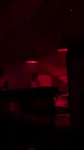 The scene is dimly lit, with a deep red hue permeating the atmosphere. A person stands behind a raised DJ booth, seemingly performing music. The individual is partially silhouetted, making it difficult to discern specific features but a baseball cap is visible. There are other figures, also mostly silhouetted, present in the dimly lit space. Above, a few red lights hang, providing the primary source of illumination and contributing to the dramatic ambiance. The setting suggests a nightclub or concert venue.