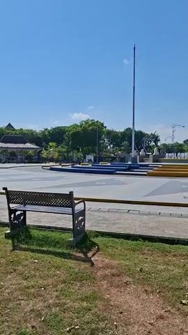 A wide shot shows a park on a clear day. In the foreground, a dark metal park bench sits on a patch of grass with dry leaves. A dirt path leads away from the bench towards a paved area. In the midground, a large, paved plaza with blue and yellow geometric patterns is visible. A tall flagpole stands in the center of the plaza, along with several statues and monuments. The largest monument features two tall, cream-colored obelisks topped with orange spires. In the background, lush green trees and a building with a brown roof are visible. The sky is a bright, clear blue.