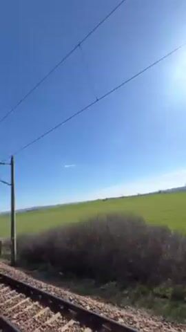 The video shows a view from a moving train. Initially, the camera faces forward, showing the train tracks, the surrounding landscape, a bright blue sky, and overhead power lines. The landscape is mostly green fields with scattered bushes. Then, the camera angle shifts to the side, revealing a window of the train. A woman and a man are leaning out of the window, smiling and posing for the camera. The woman gives a peace sign. The train continues moving, showing the tracks receding into the distance and the green landscape passing by.
