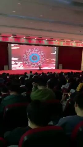 A crowd of people is seated in a large auditorium, all facing a stage. The audience members are mostly young, with some using their phones. On the stage, a person is performing in front of a large screen displaying colorful graphics. The backdrop changes periodically. The auditorium has rows of red seats and overhead lighting. There is a banner on the stage, with Chinese characters displayed.