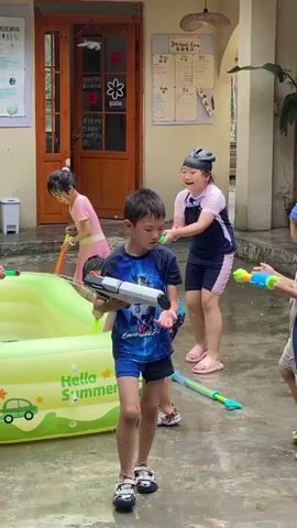 The video depicts several children engaged in a water fight. In the foreground, a young boy in a blue and black swimsuit stands holding a large white water gun. Behind him, other children are playing in and around a partially inflated green kiddie pool with "Hello Summer" printed on it. One girl in a pink and navy swimsuit smiles as she holds a pink water gun. Another boy in a blue cap and blue and grey swimsuit crouches near the pool, aiming a yellow and green water gun. A girl in a purple dress with a white cap is reaching into the pool. Other children are visible in the background, also participating in the water fight with water guns. The environment appears to be an outdoor courtyard or paved area, with a building entrance visible in the background and some plants. Water is visibly sprayed in the air, and the ground is wet.