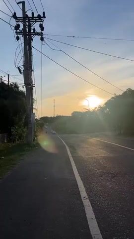 The video shows a road with vehicles moving along it. The sun is visible in the sky, suggesting it is either sunrise or sunset. There are power lines and utility poles on the side of the road, as well as trees and vegetation. The road itself appears to be asphalt and has some potholes.
