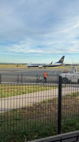 The video shows an airport tarmac with a Ryanair airplane in the background. A person in an orange vest is pushing a trolley on the tarmac. There is a fence in the foreground. The sky is blue with some clouds. Three flags are visible on flagpoles to the left.