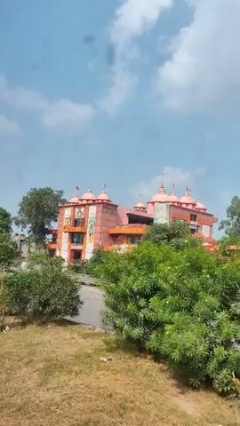 A medium shot captured from a moving vehicle shows a pink and orange building with multiple domes, possibly a temple, situated behind a road and some trees. The sky is partly cloudy. The building has several windows and balconies, and what appear to be murals or paintings on its facade. The foreground is filled with lush green foliage, obscuring the lower portion of the scene. The camera pans slightly from left to right, offering a different perspective of the building and its surroundings.