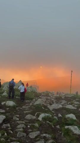 The video displays a mountainous landscape under a hazy, orange sky, possibly at sunrise or sunset.  Several people are visible, some standing on rocky terrain, others appearing to be on a platform or structure in the middle ground.  The sun is a small, bright circle in the hazy sky.  One person in the foreground, wearing a backpack, faces away from the camera. Another person in the foreground, wearing a white top and dark pants, also faces away.  The ground is covered with rocks and some patches of green vegetation.  A yellow marker post with a red top is visible on the right side of the frame.