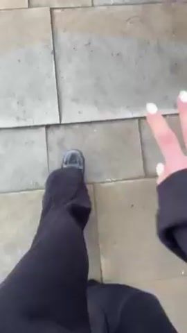 A person with pale skin and white nail polish on their fingernails holds their hand up in front of them, curling it into a fist, then opening their fingers, palm-out, and then dropping the hand down. The person wears a black long-sleeved garment. The background is a paved sidewalk made of large square tiles.