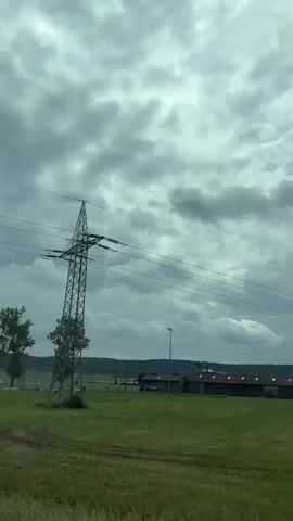 The video captures a moving perspective of a rural landscape under a heavily overcast sky. Initially, a prominent, tall lattice-style electricity pylon with multiple power lines dominates the foreground against the cloudy backdrop. Green grassy fields extend around the pylon, with distant buildings and what appears to be a sports field in the background.

As the camera continues to move, a wire mesh fence comes into view, revealing a sports area. A sand court equipped with a yellow net, typical for volleyball or beach volleyball, is visible behind the fence. Beyond this court, expansive green fields suggest soccer pitches, with white goalposts discernible in the distance. Several large, leafy trees are interspersed throughout the landscape, framing the view.

Further into the background, scattered buildings with dark roofs, possibly agricultural or industrial, are seen near a treelined horizon. A street lamp is also visible near the sports fields. The overall scene depicts a functional, semi-rural environment in Bad Windsheim, Germany, characterized by utility infrastructure, sports facilities, and natural elements like trees and fields, all under a dull, cloudy daytime sky. No people or visible text are present in the footage.