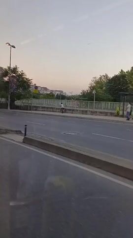A video recorded from a moving vehicle shows a street scene with a bus stop. Several people are visible at the bus stop, some standing and some sitting. A person on a bicycle rides by on the sidewalk. In the background, there are trees, buildings, and a minaret of a mosque. A black van drives past the camera. The sky is visible and appears to be clear.