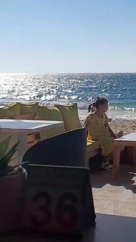 A video shows a young child sitting on a couch at an outdoor cafe by the sea. The child is wearing a yellow dress. There is another child to the left of the first child. The sea is visible in the background with waves and sunlight reflecting on the water. The cafe has tables and chairs. A person wearing a black cloak and a hat walks into the frame from the left.