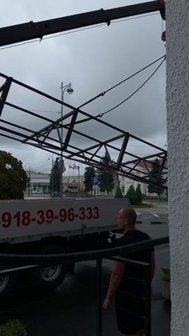 A crane is lifting a metal structure. A truck with the number 8-918-39-96-333 is parked below. A man in a black t-shirt is walking away from the truck. The sky is cloudy.