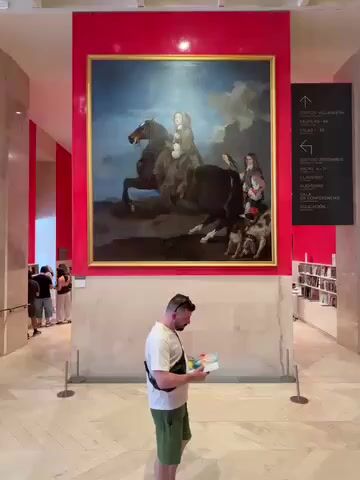The image displays the interior of a spacious art museum or gallery, located in Madrid, Spain, characterized by modern architecture and bright lighting. In the foreground, a man in a white t-shirt, green shorts, and a black crossbody bag stands with his back to the camera, intently reading from a small book or guide.

He is positioned before a massive oil painting, centrally displayed on a prominent, glossy red wall panel. The painting itself depicts a dynamic equestrian scene under a cloudy blue sky. Two main figures are shown on horseback: one, appearing younger, on a lighter-colored horse, and another adult figure on a darker horse. Two dogs accompany the adult's horse. The painting is set in a golden frame.

To the left in the background, several other museum visitors are visible, engaged in viewing exhibits or walking through the space. On the right, a tall black directional sign with white text provides guidance within the museum, listing areas such as "EDIFICIO VILLANUEVA" (Villanueva Building), "SALAS 00-60" (Rooms 00-60), "EDIFICIO JERÓNIMOS" (Jerónimos Building), "CLAUSTRO" (Cloister), "AUDITORIO" (Auditorium), "SALA DE CONFERENCIAS" (Conference Room), and "EDUCACIÓN" (Education), accompanied by directional arrows. Further to the right, shelves stocked with books or merchandise can be seen. The museum features light-colored tiled floors and clean white walls, with metal stanchions cordoning off the area around the large painting, indicating it's a significant exhibit. The scene captures a typical daytime visit to an art institution.
