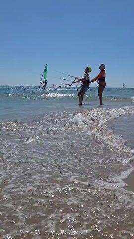 Two people stand in shallow ocean water near a sandy beach. The person on the left is wearing a dark shirt, denim shorts, and a yellow helmet. They are holding onto a kiteboarding control bar, with two lines extending upwards and to the left. The person on the right, wearing an orange life vest and a hat, has their hands on the back of the person on the left. They appear to be instructing or assisting. In the background, several other individuals are windsurfing on the ocean, with sails of various colors visible. The water is choppy with waves breaking on the shore. The sky is clear and blue.