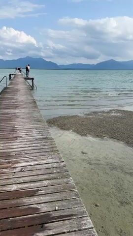 A drone shot pans across a wooden pier extending into a calm lake. Two people stand at the end of the pier, looking out at the water. The lake stretches to the horizon, where a range of mountains forms a dramatic backdrop. The sky is partly cloudy, with patches of blue sky visible between the white clouds. The water is a clear, turquoise color, with gentle waves lapping at the shore. The pier is made of weathered wooden planks, with a railing along one side. The shore is made up of pebbles and sand. The scene is peaceful and serene, evoking a sense of tranquility and natural beauty.