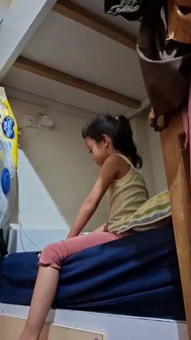 A young girl with dark hair pulled into a ponytail, wearing a light yellow tank top and pink leggings, sits on a dark blue mattress, possibly a bunk bed. She is positioned on the left side of the frame and her right arm is extended forward, her hand blurred from movement, as if she is gesturing or pointing. To her left, a white and blue oscillating fan is visible. Behind her, a wooden structure, likely part of a bunk bed, is present, with some fabric hanging on the right side. The wall behind the girl appears to be beige. The lighting suggests an indoor setting, possibly a bedroom.