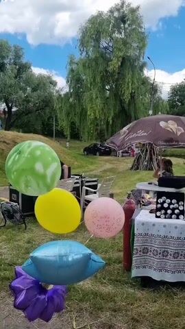 The video shows a cluster of balloons floating in an outdoor setting. There are green, yellow, pink, purple, and blue balloons. In the background, there are several white chairs, sound equipment, a table with a patterned tablecloth displaying small items, and a large umbrella. A person is seated in front of the sound equipment, and another person is visible behind the table. The environment appears to be a park or a grassy area with trees and a cloudy sky.
