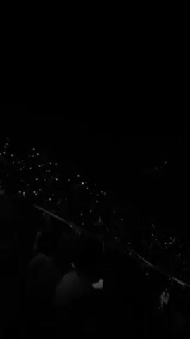 The video shows a concert in a dark arena. The stage is dimly lit and adorned with concentric arches. A large screen displays a man singing into a microphone. The audience, barely visible in the darkness, holds up glowing phones, creating a field of lights.