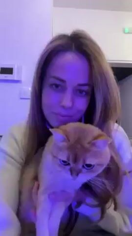A young woman with light brown hair is holding a tan cat. She is sitting in a room with white walls and a partially open white door. There is an electrical panel on the wall behind her. She puckers her lips at the cat. There is music playing.