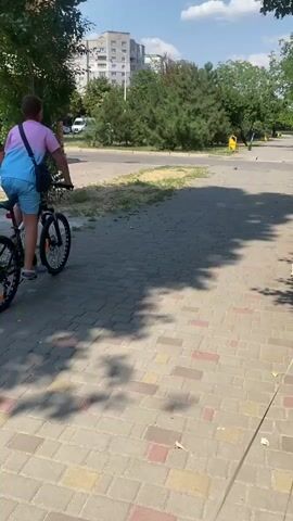A person rides a bicycle down a paved path on a sunny day. The person is wearing a pink and blue tie-dye t-shirt and blue shorts. They are carrying a blue bag over their left shoulder. The path is made of brick pavers and is lined with grass and trees. In the background, there are apartment buildings and more trees. The sky is blue with some scattered clouds. The video is shot from behind the person on the bicycle, following them as they ride away from the camera.