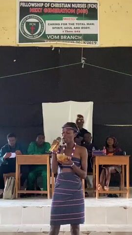 This media file depicts an indoor event, likely a gathering or performance, taking place during the daytime. The setting appears to be a room with a stage area at the front, indicated by a raised platform and a backdrop of black fabric.

**Subjects:**
The central figure is a young woman dressed in traditional attire, including a patterned skirt and a beaded necklace. She is holding a microphone and a bowl, seemingly addressing an audience. Behind her, several other individuals are seated at tables. These include men and women, some appearing to be participants or officials, and at least one younger person. One person is wearing a red cap.

**Objects:**
Visible objects include microphones, wooden tables, chairs, a bowl, a backpack, papers, and what appear to be bottles or containers on one of the tables. A banner is prominently displayed at the top of the scene.

**Activity or Event:**
The main activity is a presentation or performance by the young woman. She is holding a microphone and a bowl, suggesting she is speaking, singing, or perhaps part of a dramatic presentation. The presence of seated individuals at tables indicates an audience or judging panel.

**Notable Details:**
The lighting suggests it is daytime. The room has a somewhat utilitarian appearance, with simple furniture and fabric backdrops. The clothing of the central figure and some of the audience members suggest a cultural or traditional context. The mood appears to be one of focused attention on the speaker.

**Visible Text:**
The banner at the top reads:
"FELLOWSHIP OF CHRISTIAN NURSES (FCN)
NEXT GENERATION (NG)

MOTTO:
CARING FOR THE TOTAL MAN;
SPIRIT, SOUL AND BODY.
1 Thess. 5:23

VOM BRANCH
20/8/2019"