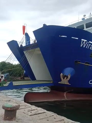 The video shows a large blue and white ferry named "Wira Loewisa" docked at a pier. The ferry is tied to the pier with a thick yellow rope. The sky is overcast, and the water is calm. In the background, a large industrial plant with tall structures can be seen. The ferry is viewed from a close-up perspective, focusing on its side and bow.