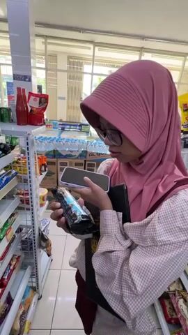 A young woman wearing a pink hijab and glasses stands in a convenience store aisle. She holds a phone in her left hand and a chocolate bar in her right. The phone is positioned as if recording a video. She appears to be speaking to the phone, possibly reviewing the chocolate bar. Shelves stocked with drinks and other products are visible in the background. The lighting suggests an indoor environment, possibly during the day.